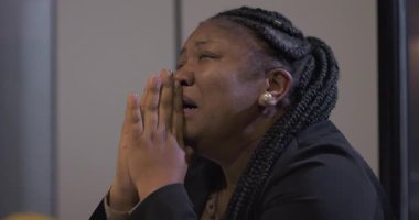 Afro-Amerikalı iş kadını, ofis masasında oturuyor, yüzü ellerine gömülü, sessizce dua ediyor ve ezici iş baskısı yüzünden duygusal olarak yorgun düşüyor.