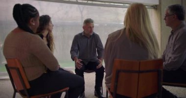 Duygusal grup terapisinde kişisel mücadeleyi ortaya koyan, katılımcılar güvenli iyileşme alanında empatiyle dinlerken, aciliyet ve kırılganlıkla konuşan bir adam.