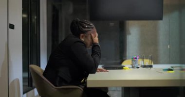 Afrikalı Amerikalı iş kadını, uzun ve yoğun bir iş gününden sonra, duygusal stres ve yorgunluğu aşmak için, elleri yüzü kapalı bir şekilde ofis masasında sessizce oturuyor.