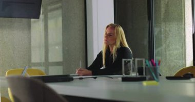 Kadın, modern ofis ortamında masa aksesuarları, dizüstü bilgisayarlar ve sandalyelerle çevrili, düşünceli bir şekilde konferans masasında oturuyor.