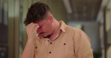 Ofis koridorunda el ele tutuşan bir adam, hayal kırıklığını ve stresi doğal ışıkla sessiz profesyonel çalışma alanında gösteriyor.