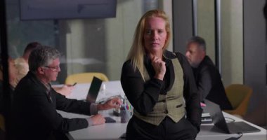 Ofisteki takım toplantısında gergin ve derin düşüncelere dalmış sarışın profesyonel bir kadın. İş arkadaşları işlerine odaklanırken masanın yanında duruyor.