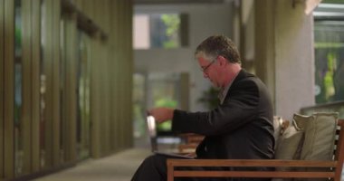 Ofiste oturan orta yaşlı bir adam dizüstü bilgisayarı açıyor ve takım elbisesini ayarlıyor, doğal ışık ile sessiz profesyonel çalışma alanında çalışmaya hazırlanıyor.