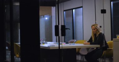 Orta yaşlı iş kadını boş konferans masasında tek başına otururken, modern ofis alanındaki zor şirket toplantısından sonra yansıtıcı ve stresli bir görünüm sergiliyor.