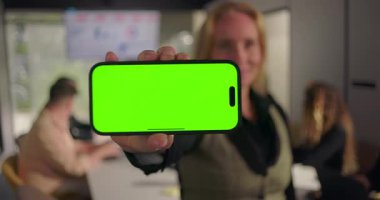 Yakın plan, yeşil ekranlı akıllı telefon, bulanık iş adamları modern konferans odasında ekranda çizelgelerle çalışıyorlar.