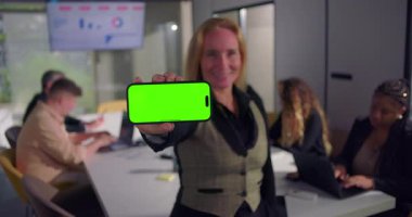 Modern ofiste elinde yeşil ekranlı akıllı telefonu olan gülümseyen iş kadını, arka planda dizüstü bilgisayarlar ve çizelgeler üzerinde çalışan meslektaşları