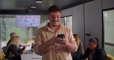 Toplantı sırasında akıllı telefona gülümseyen genç işadamı, meslektaşları modern konferans odasında veri ekranı ile dizüstü bilgisayarlarda çalışırken mutlu tepkiler verdi