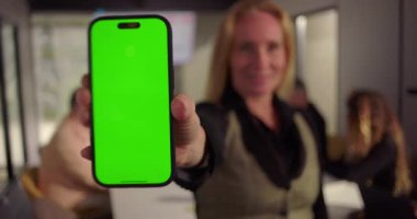 Ofis toplantısında akıllı telefondan yeşil ekran sunan iş kadınının yakın çekimi. Çeşitli takımlar konferans masasında çalışıyor. Analizler ekranda.