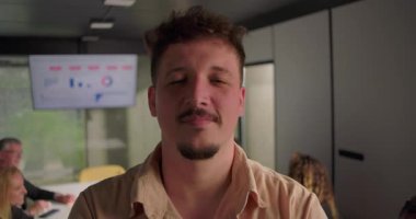 Ofisteki kameraya bakan ciddi genç adamın yakın çekimi, arka planda çalışan meslektaşlar büyük ekrandaki dijital veri tablosunun yanında