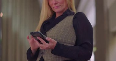 İş kadını, iş günü ofis koridorunda dikilirken sakin bir şekilde akıllı telefona bakıyor.