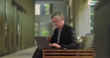 Orta yaşlı bir adam sessiz ofis salonunda dizüstü bilgisayar kullanıyor, modern kapalı çalışma alanında doğal ışıkla çalışırken ekrana odaklanıyor.