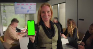 Ofis toplantısında elinde yeşil ekranla kameraya doğru gülümseyen iş kadını, profesyonel ortamdaki dijital ürün yerleştirme fırsatını gösteriyor