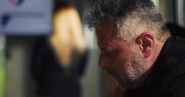 Üzgün işadamı, ofisteki çatışmalardan sonra sessizce aşağıya bakıyor, duygusal olarak etkilenmiş ve soyutlanmış, kadın meslektaşı ise arka planda duruyor, işyeri gerilimi ve pişmanlığı.