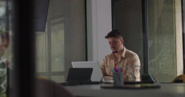 Genç erkek profesyonel, sessiz ofis günlerinde dizüstü bilgisayarda tek başına çalışır. Modern masadaki doğal ışık ve minimum dikkat dağınıklığıyla görevine odaklanır.