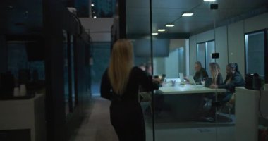 Meslektaşlar modern kurumsal çalışma ortamında geç saatlerde ofis toplantılarında dizüstü bilgisayarlarla masaya otururken cam toplantı odasına giren kadın yönetici.