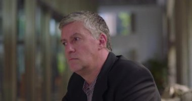 Ofiste oturan orta yaşlı bir adam düşünceli bir ifadeyle mesafeye bakıyor. Modern profesyonel çalışma alanına sessizce yansıyor.