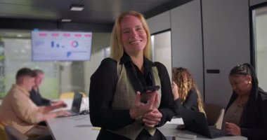 Gülümseyen iş kadını elinde akıllı telefon, arka planda veri tabloları olan dizüstü bilgisayarlar üzerinde çalışan çeşitli meslektaşları olan toplantı odasında duruyor.