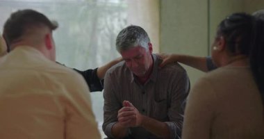 Adam grup terapisi sırasında başını eğiyor ve ellerini tokuşturuyor. Etrafı sakin ve şefkatli ellerle destek veren akranlarıyla çevrili.