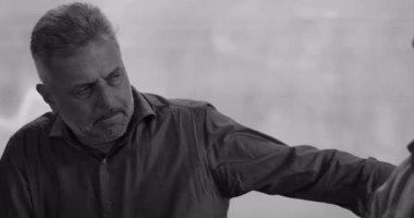 Grup terapisi sırasında çöken bir adam, acı içinde başını eğerken başka bir grup üyesi omzunda rahatlatıcı bir elle sessiz destek sunuyor, siyah ve beyaz.