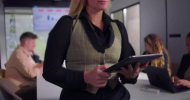 Ofis toplantısını yöneten kendine güvenen iş kadını, tablet hakkındaki bilgileri incelerken, arka planda çalışan destekleyici ve odaklı ekip, modern liderlik tarzını vurguladı