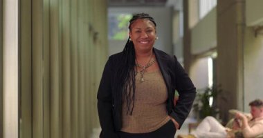 Modern ofis koridorunda el ele tutuşmuş gülümseyen Afrikalı Amerikalı kadın rahat ve kendinden emin bir şekilde arka planda dinleniyor.