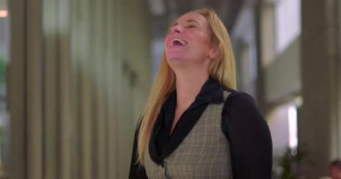 İş kadını modern ofis koridorunda kendiliğinden gülüyor, gerçek neşeli bir ifade, başı arkaya eğik ve gözleri kapalı.