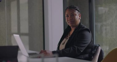 Kendine güvenen Afrikalı Amerikalı iş kadını dizüstü bilgisayarda oturuyor ve şirket ofisinde göz teması kuruyor.