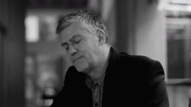 Yaşlı bir adam, kapalı gözleri ve kederli ifadesiyle hafifçe arkasını dönüyor, evde otururken stres veya duygusal bunalımı tasvir ediyor.