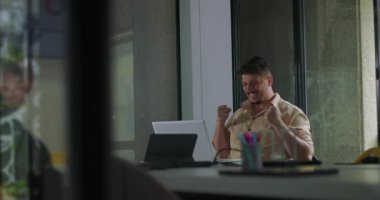 Adam başarıyı ofis masasında heyecanlı kol hareketiyle kutluyor. Modern iş yerinde olumlu sonuçlar aldıktan sonra dizüstü bilgisayara gülümsüyor.