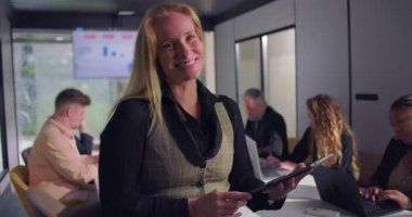 Ofis toplantısı sırasında konferans masasının başında duran neşeli kadın yönetici, verimli ve olumlu bir ortamda arka planda çalışan takım.