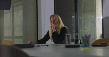 Kadın tek başına konferans masasında oturuyor, yüz yüze, düşünceli ya da stresli görünüyor, modern ofis ortamında cam duvarlar ve masa aksesuarları görünür