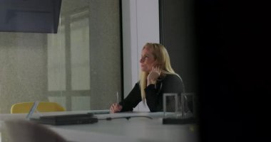 Konferans masasında oturan kadın, modern ofiste el ele tutuşarak, gün ışığında ve işyerinde derin derin düşüncelere dalmış ve odaklanmış görünüyor.