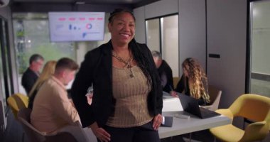 Gülümseyen Afrikalı Amerikalı iş kadını konferans masasının yanında duruyor. Arka plandaki toplantıda çalışan iş arkadaşlarıyla kendinden emin pozlar veriyor.