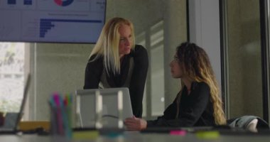 Açıklamak için masaya yaslanan sarışın iş kadını, ofiste yakın işbirliği sırasında yanında veri tablosuyla oturan meslektaşını işaret ediyor.