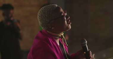 Evanjelik vaiz tutkulu bir şekilde cemaate hitap ediyor, yakın profil, gümüş saçlı Afro-soylu, gözlüklü, pembe ceketli, güçlü vaaz veren bir kadın.