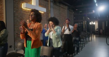 Cemaat kilise ayini sırasında el çırpıyor, arka planda haç ve tuğla duvarlar aydınlatılıyor, çeşitli topluluklar neşe ve birlik içinde.
