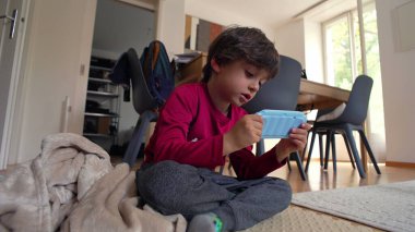 Yerde oturan genç bir çocuk, iki eliyle bir oyuncak kamera tutuyor, ev ortamında oyalanırken dikkatle odaklanıyor.