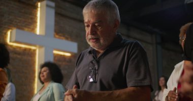 Kilisede namaz kılarken yansıyan yaşlı adamın yakın çekimi, sadakat içinde sıkıca sarılmış eller, arka planda aydınlatılmış haç, derin inanç ve ruhaniliğin ifadesi.