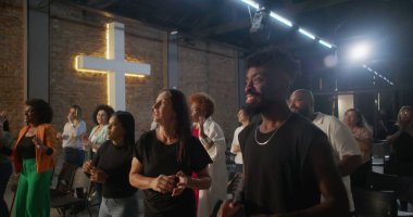 Kiliseye ibadet sırasında alkış tutan ve gülümseyen çok kültürlü cemaat, arka planda aydınlatılmış haç, inanç, neşe, birlik ve ruhani bağlılık ifadeleri.