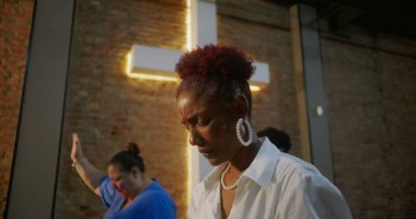 Kilisenin içinde dua eden bir kadın, minnettarlığını ve inancını ifade eden, arka planda diğer ibadet edenlerle birlikte ışıldayan bir haçın önünde duran.