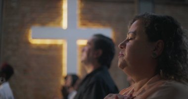 Çeşitli grup ibadetleri sırasında elleri kalbimde olan kadın, arka planda aydınlatılmış haç, maneviyat, inanç ve minnettarlık vurgulandı