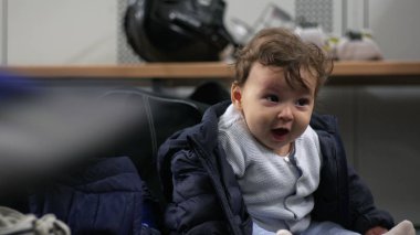 Bebek soyunma odasında oturuyor, heyecandan gülümsüyor, sıcak kıyafetlerle sarmalanmış, neşeli ve canlı bir an..