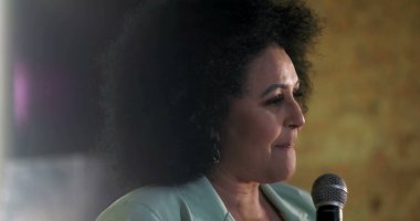 Ruhani bir olay sırasında mikrofona tutkulu bir şekilde konuşan bir kadının görüntüsü. Kendine güvenen liderlik, duygusal ifade ve izleyicilerle iletişim.