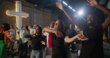 İbadet sırasında sevinçle el kaldıran ve dans eden farklı bireylerden oluşan bir grup, arka planda aydınlanan haç, canlı bir manevi bağlantı anı ve kutlama anı.