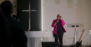 Vaaz veren, mikrofon tutan, arka planda haç olan kilisedeki cemaate hitap eden, dinleyicilerin el çırpması, Hıristiyan ruhani buluşması.