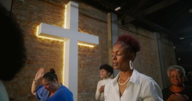 Kilisenin içinde dua eden bir kadın, minnettarlığını ve inancını ifade eden, arka planda diğer ibadet edenlerle birlikte ışıldayan bir haçın önünde duran.