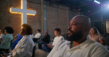 Kilisedeki sandalyeden kalkan, ellerini göğsüne koyan, dua ederken gözlerini kapayan, arka planda ışık saçan, topluma tapınan ve ruhani yansıması olan bir adam.