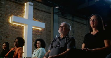 Arka planda aydınlanmış haçla kilisede oturan çeşitli insan grupları, ibadet sırasında maneviyat ve sadakat, birlik ve dini cemaatle bağlantıyı ifade ediyorlar.