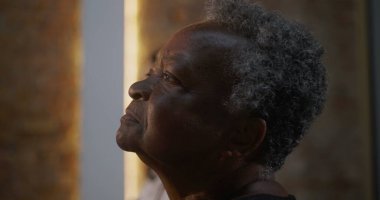 Kilisede ibadet ederken sakin bir ifadeyle profil görünümlü yaşlı bir kadın, arka planda aydınlanmış haç, maneviyat, inanç ve odak noktasında toplum