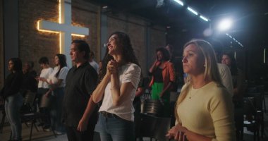 Arka planda parlayan bir haçla Hıristiyan kilisesi etkinliğine katılan çeşitli insanlar ellerini kaldırarak ibadet ediyorlar.